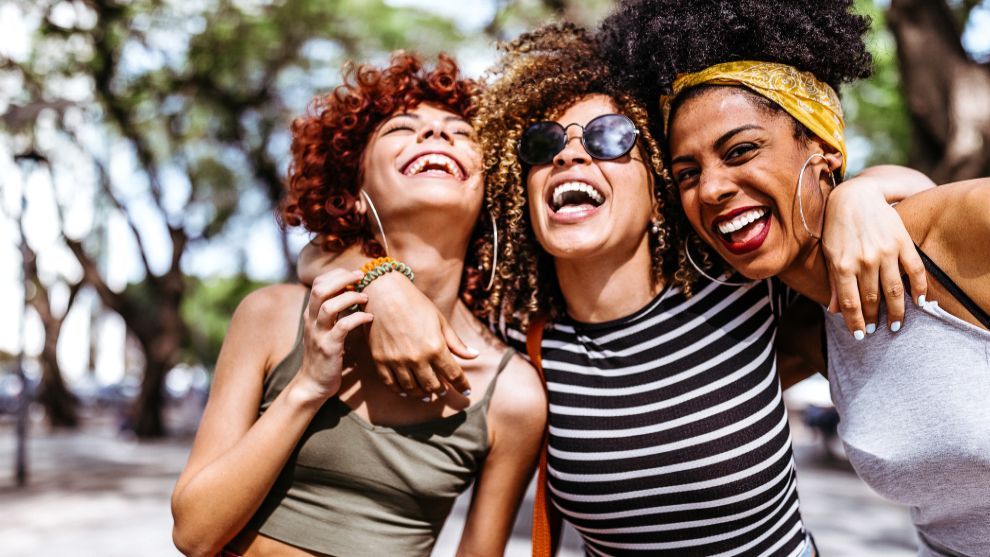 Drei junge, lachende Frauen mit lockigen Haaren, die sich umarmen.