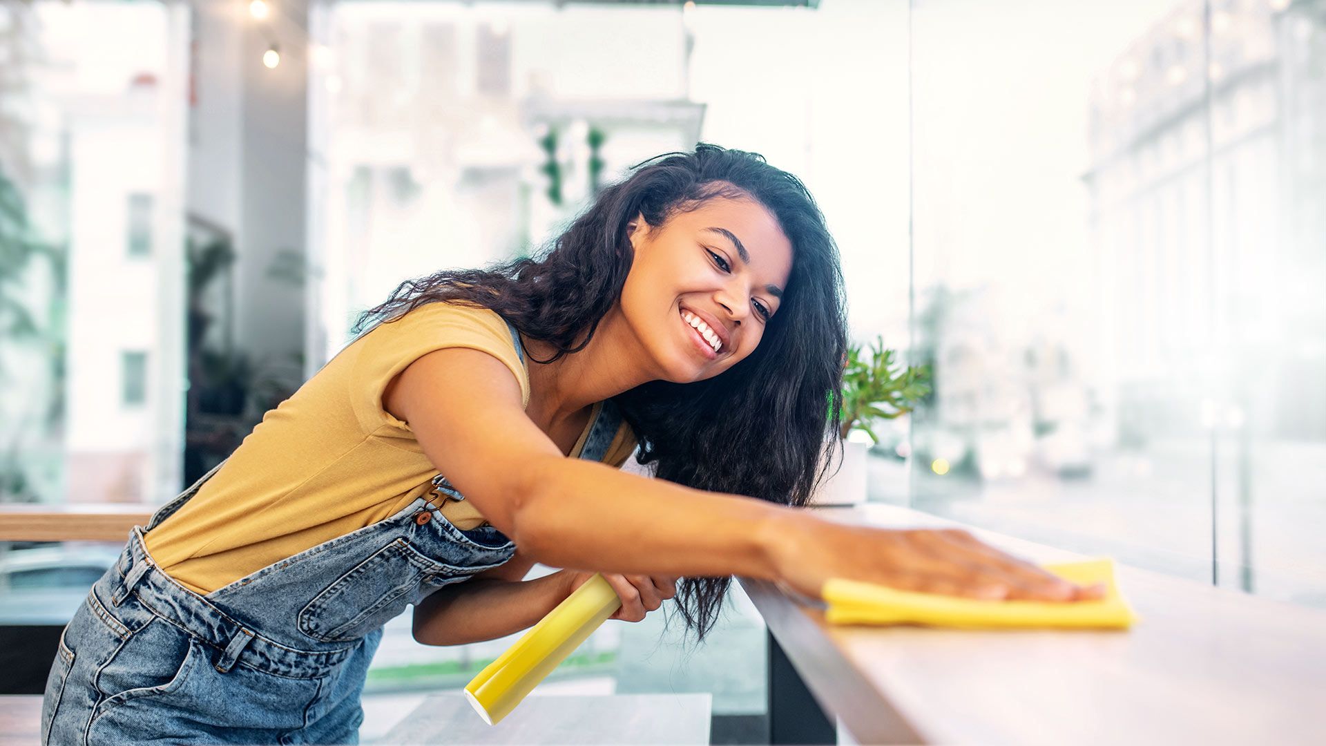 Eine junge Frau im Overall lächelt, während sie in einem hell erleuchteten Raum mit großen Fenstern eine Oberfläche mit einem gelben Tuch reinigt.