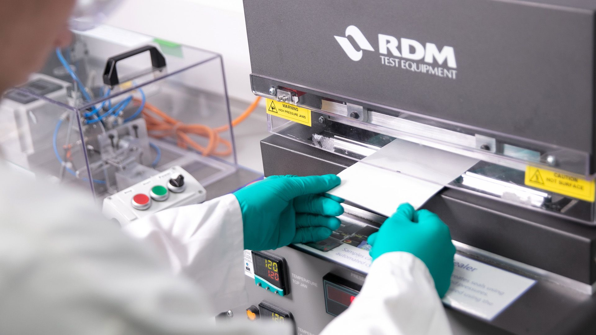 Ein Wissenschaftler aus der Forschung und Entwicklung von Unilever führt in weißem Laborkittel und grünen Handschuhen eine Papierverpackungsprobe in eine Maschine ein.