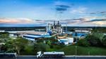 Industriegelände mit Tanks, Gebäuden und einem Tankwagen auf einer Straße vor etwas Grün und einem blauen Himmel mit Wolken.