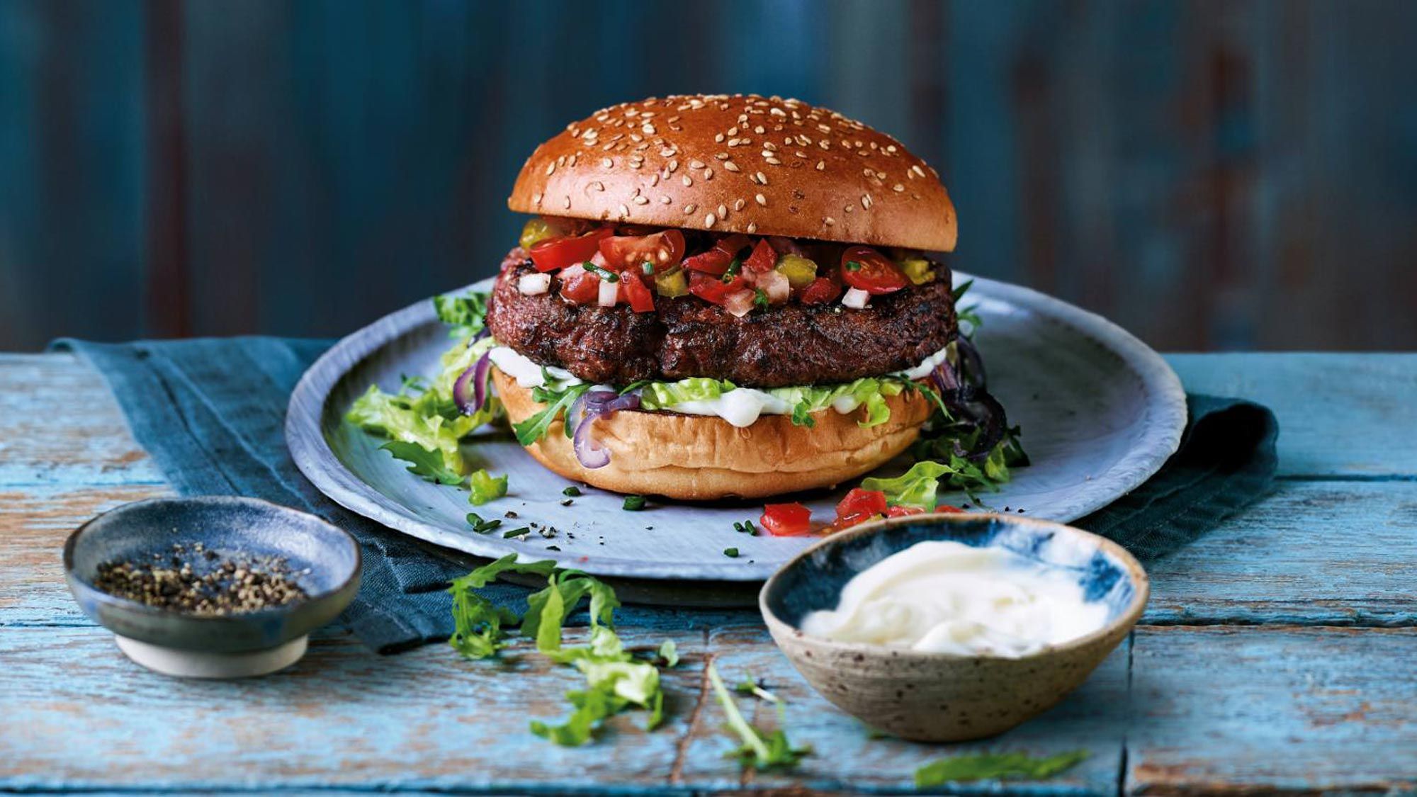 Ein Burger auf einem Teller und eine Schale mit Mayo auf einem rustikalen blauen Tisch.