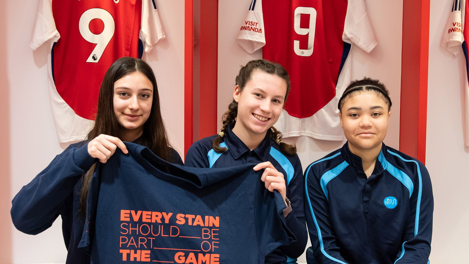 Drei Mädchen in der Umkleidekabine des Arsenal FC halten ein T-Shirt mit dem Schriftzug „Every stain should be part of the game“.
