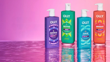 Vier farbige Flaschen von Olly Mood + Skin Waschlotion auf einer reflektierenden Oberfläche vor einem Hintergrund mit rosafarbenem Farbverlauf.